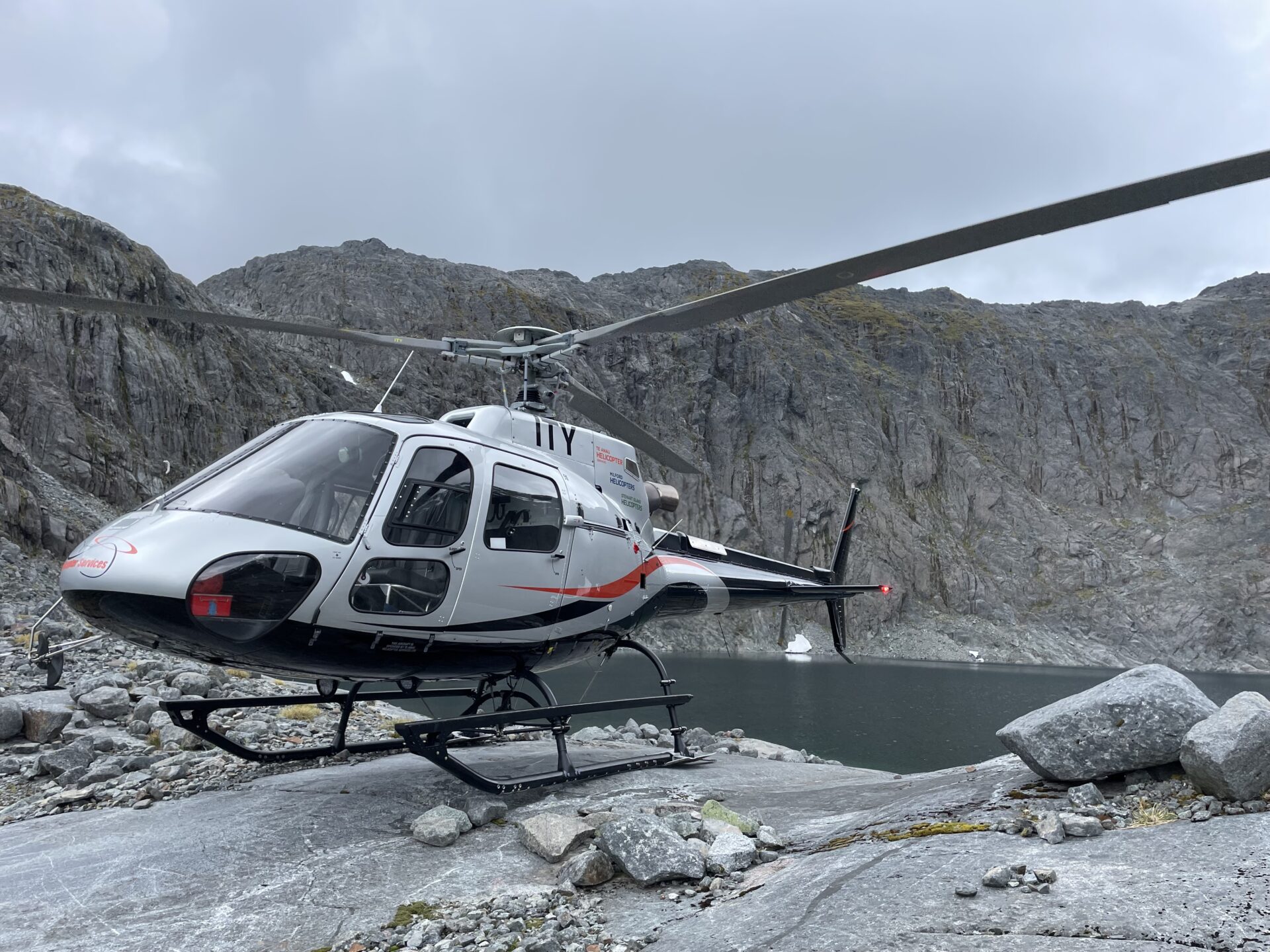 Helicopter landed on rocks in front of lake with rocky hill behind.
